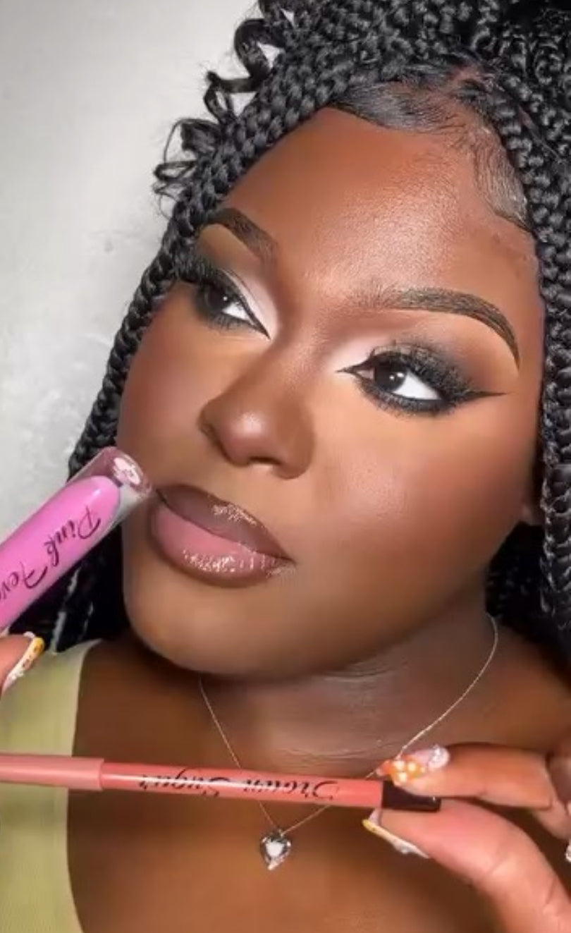 A woman with braided hair applies the Pink Status Lip Combo | Pink Gloss & Brown Lip Liner by Twicetheminks, holding the brown lip liner under her chin. She showcases bold eye makeup, glossy lips enhanced by her favorite pink gloss from the set, and wears a heart necklace.