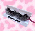 Game Over lashes feature a tapered design in a white tray printed with "Game Over," set against a pink and white patterned background. A pink applicator displays one lash, highlighting its natural yet glamorous look.