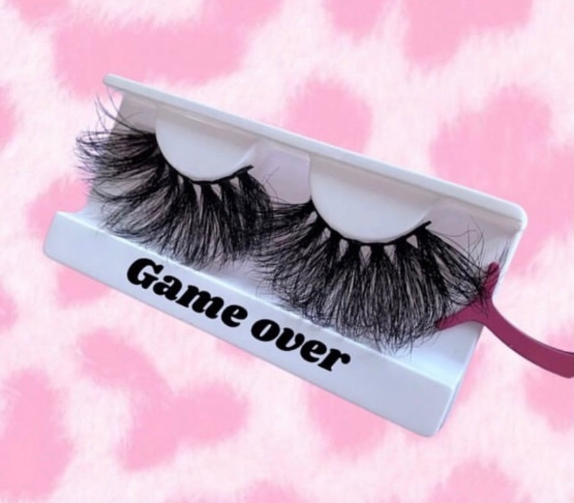 Game Over lashes feature a tapered design in a white tray printed with "Game Over," set against a pink and white patterned background. A pink applicator displays one lash, highlighting its natural yet glamorous look.