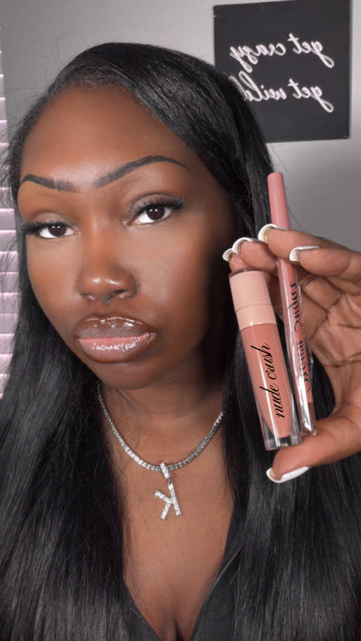 A woman with long black hair holds the Sugar Rush Lip Combo, featuring a nude-colored gloss and a matching brown sugar lip pencil. She wears a K pendant necklace and her lips are glossy and nude. In the background, a decorative sign is softly blurred.